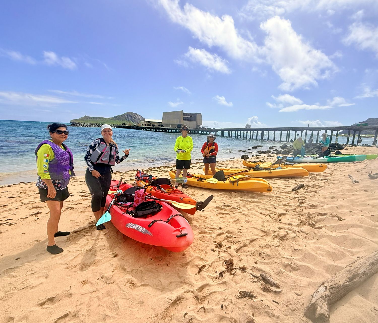 Four women posing with seven kayaks on the beach with the Makai Range Pier in the background